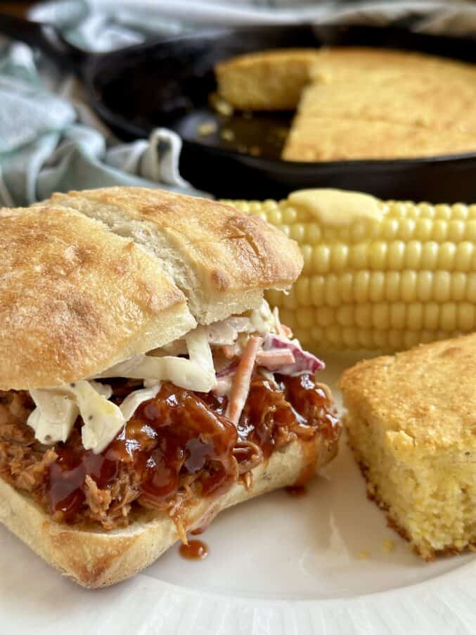 Bbq pulled pork crockpot recipe showing sandwich with cornbread and corn on the cob with pat of butter on a plate