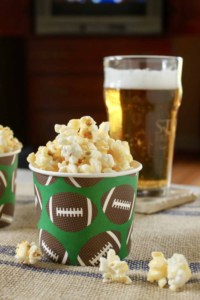 photo of caramel crunch popcorn in a paper cup with brown footballs on it with a beer behind for best tailgate party food