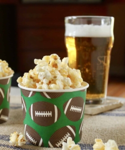 photo of caramel crunch popcorn in a paper cup with brown footballs on it with a beer behind