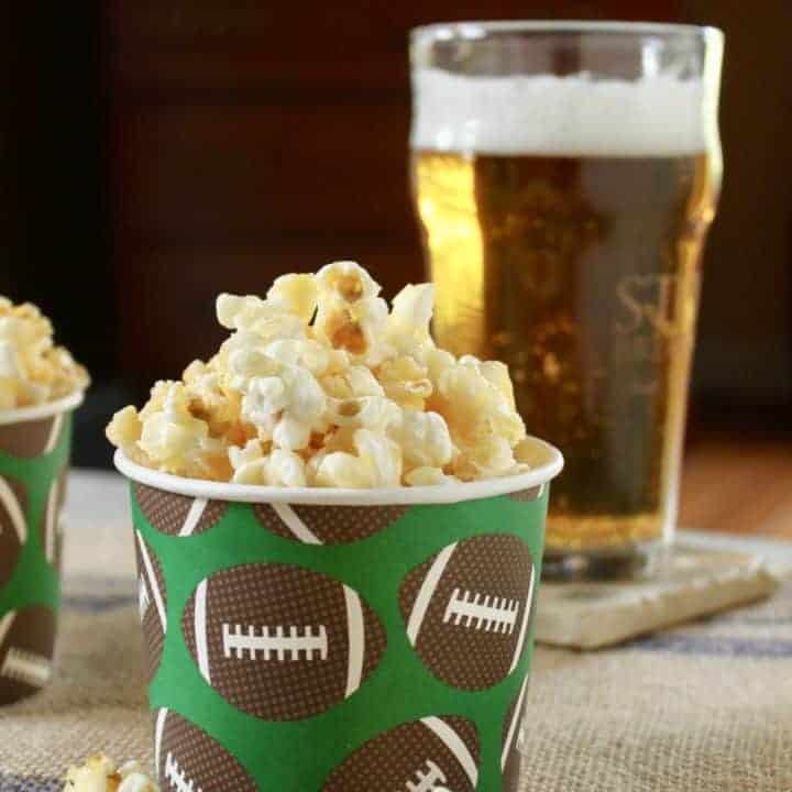 photo of caramel crunch popcorn in a paper cup with brown footballs on it with a beer behind