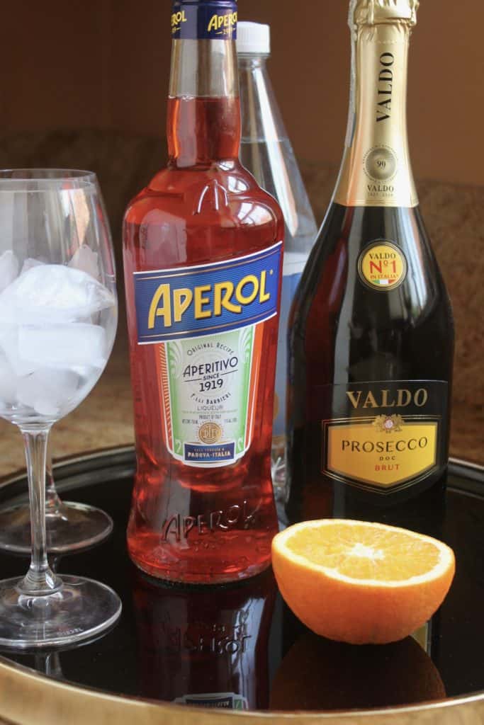 bottle of Aperol, prosecco, half an orange and glasses with ice on a round black tray