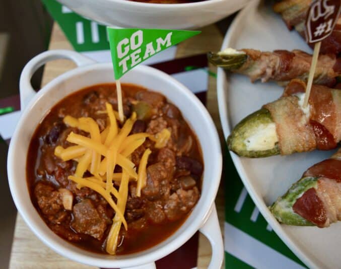 Melissa's party chili in a bowl with cheese and jalapeno poppers nearby for best tailgate party food